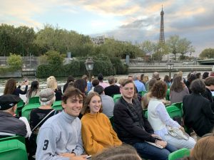 Die beiden Erlanger Studierenden und ein Pariser Kommilitone sitzen auf einem Boot und schauen in die Kamera. Um sie herum sitzen noch einige andere Personen auf dem Boot. Im Hintergrund sieht man den Eiffelturm. 