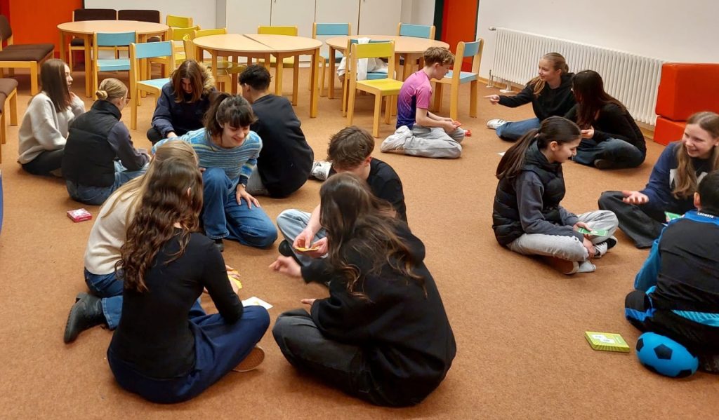 Schülerinnen und Schüler sitzen in Gruppen auf dem Boden eines Klassenzimmers und nehmen an einem interaktiven Italienisch-Workshop teil.