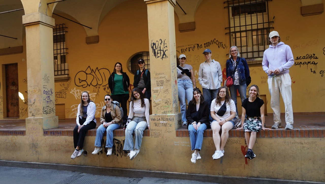 Gruppe sitzt unter Arkaden mit Graffiti an einer gelben Wand.
