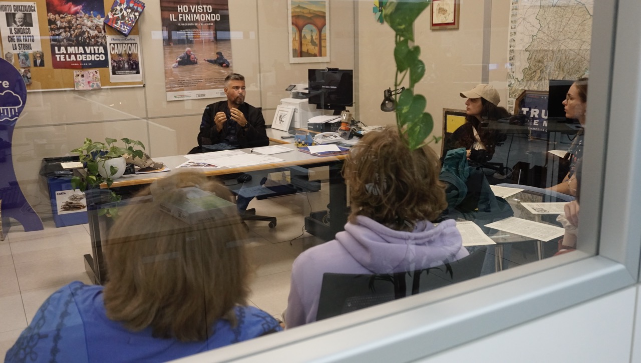 Gespräch in Redaktionsbüro; Personen um Schreibtisch, Poster an der Wand.