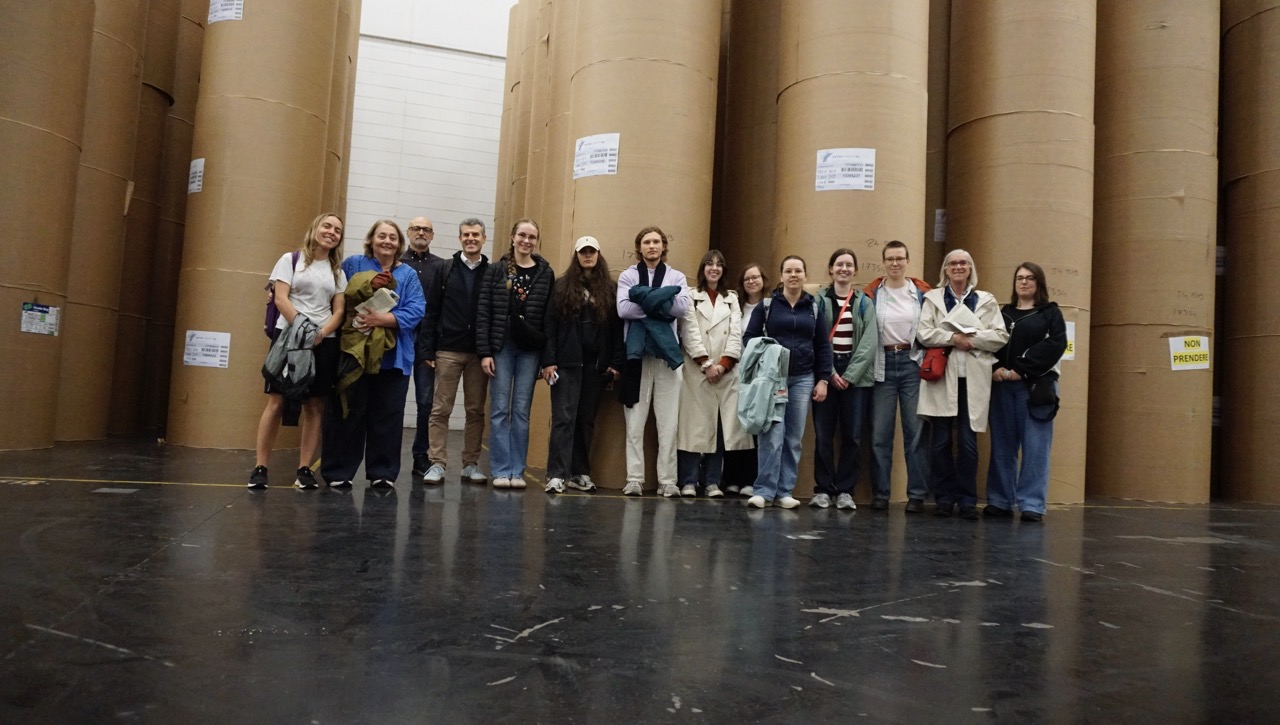 Gruppe in Druckerei vor hohen Rollen mit Zeitungspapier.