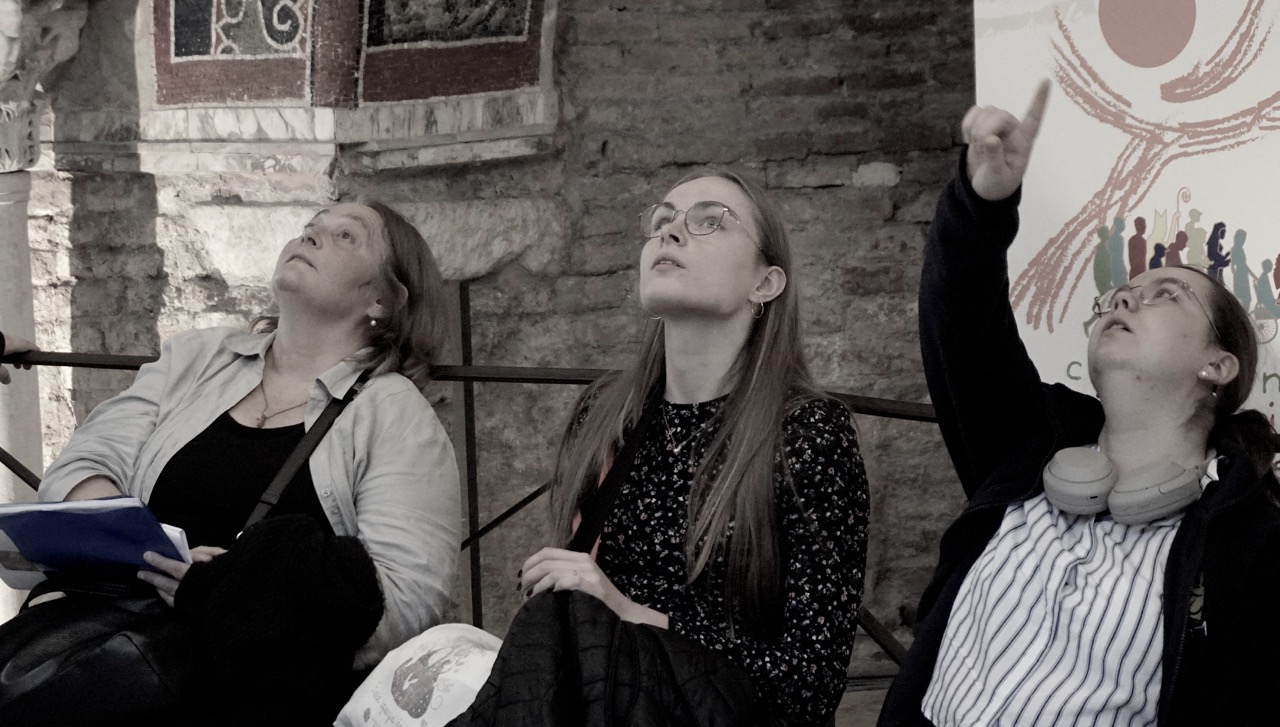 Drei Teilnehmerinnen betrachten und zeigen auf Mosaiken im Innenraum.