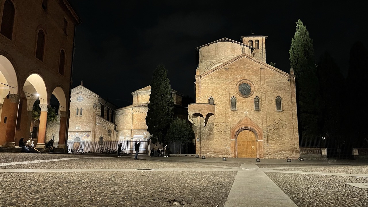 Backsteinkirche auf beleuchtetem Platz in Bologna bei Nacht, wenige Menschen im Bild.
