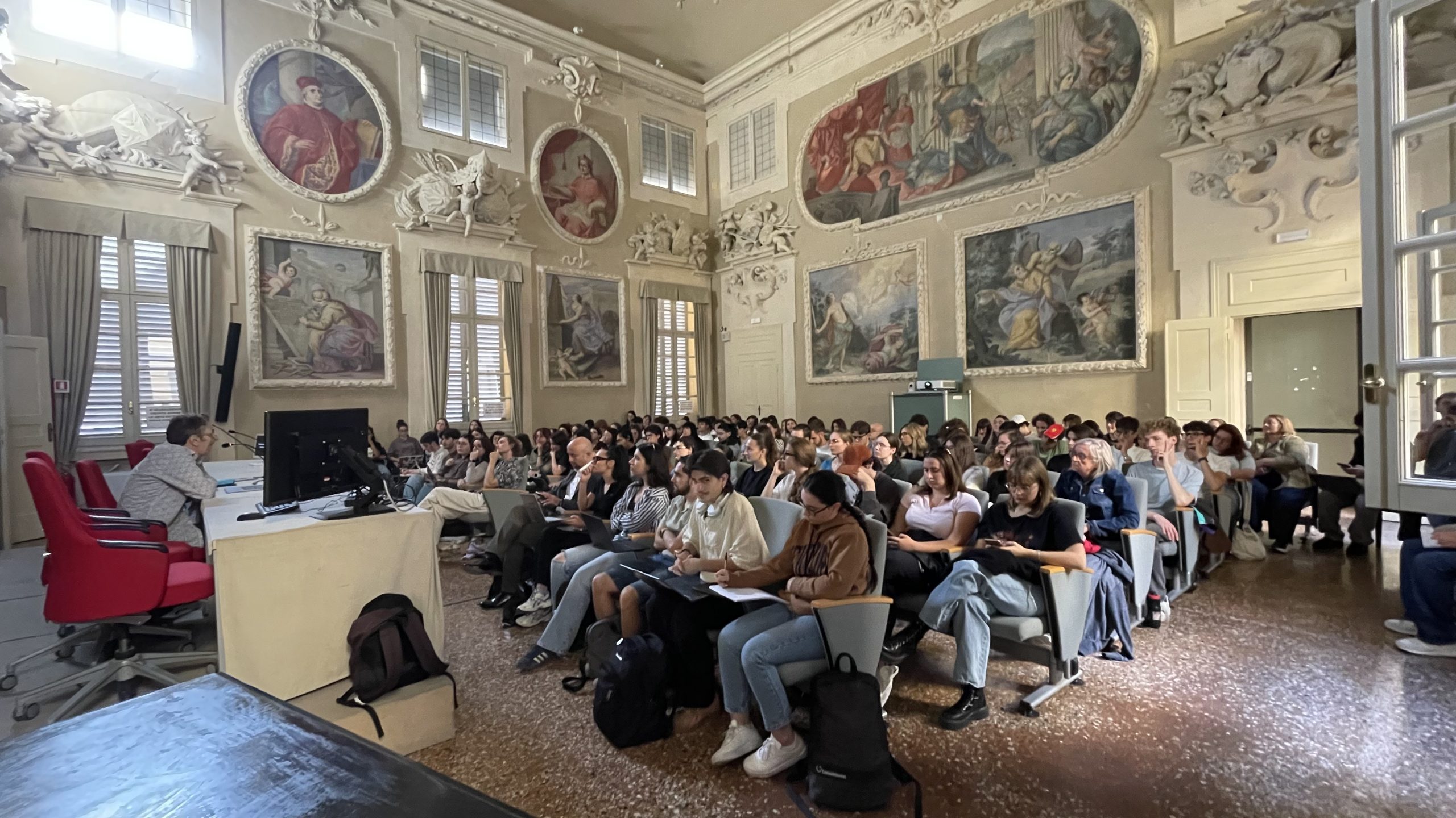 Gefüllter Uni-Hörsaal mit historischen Wandmalereien; Vortragender links.